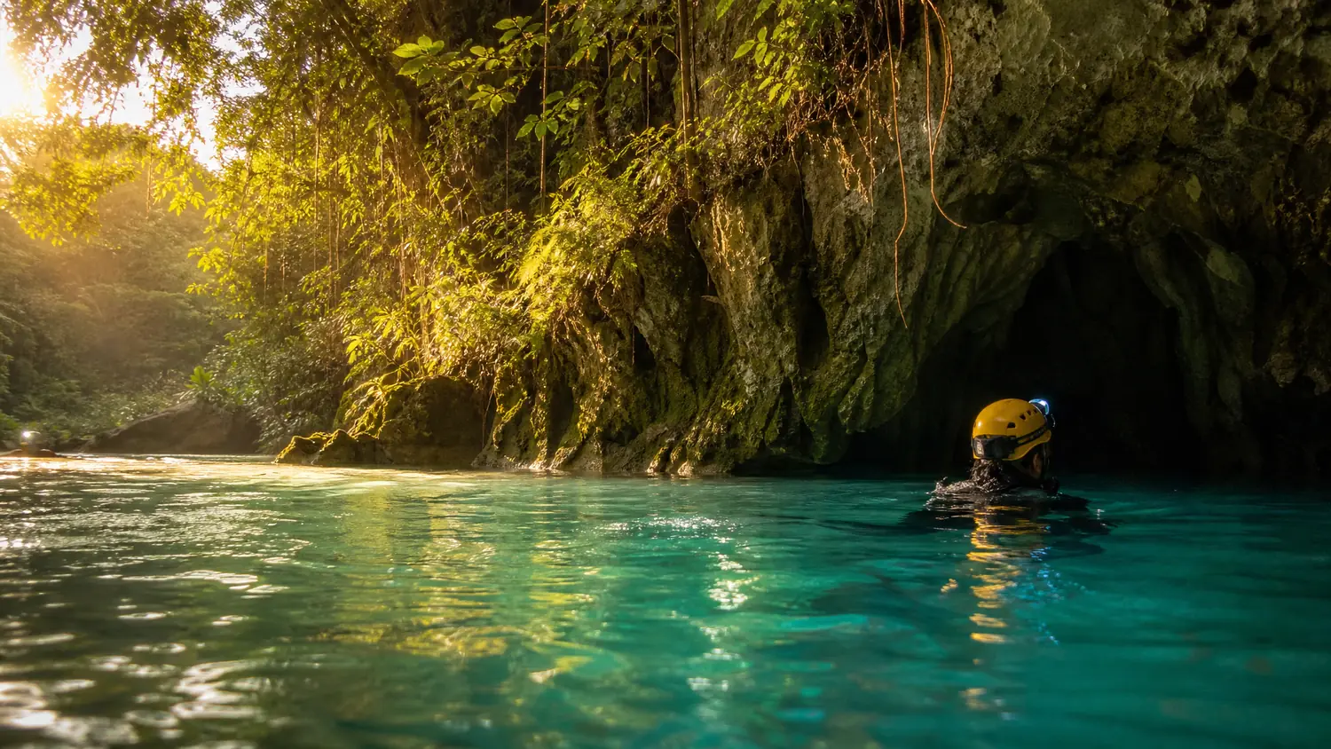 Ein Schwimmer mit gelbem Helm schwimmt im goldenen Abendlicht über grünes Wasser auf einen Höhleneingang zu