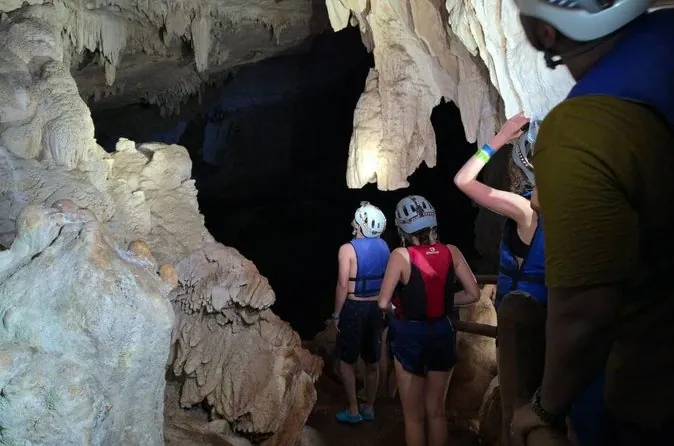 Besucher mit Helmen und Schwimmwesten, von hinten gesehen, gehen in eine Höhle mit hellen Formationen